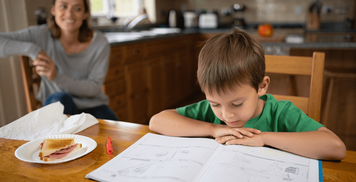 Child studying at a table with an adult in the background.